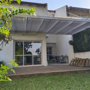 פרגולה מאלומיניום מותקנת מעל דק עץ בחצר בית.