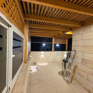 פרגולת עץ מעוצבת במרפסת מרוצפת עם חיפוי אבן ותאורת קיר