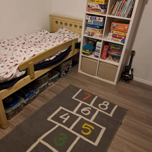 חדר ילדים עם מדף משחקים, שטיח קלאס, מיטה וארגזי אחסון.