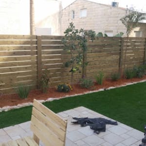 גינה עם דשא מלאכותי, צמחים ופרחים לאורך גדר עץ