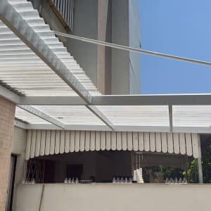 פרגולה אלומיניום לבנה בחצר עם קיר לבנים ועצים ברקע
