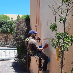 אדם צובע קיר חיצוני באמצעות רולר, תלוי בחבלים.