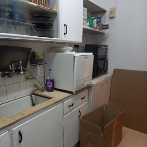 מטבח עם ארונות לבנים, קרטון פתוח, כיור וחומרי ניקוי.