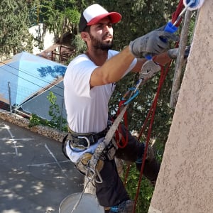 אדם צובע קיר חיצוני עם רולר, תלוי בחבלים