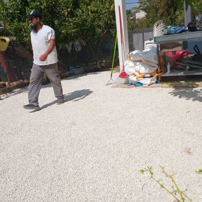 אדם הולך בחצר עם חצץ, עצים וכלי גינון