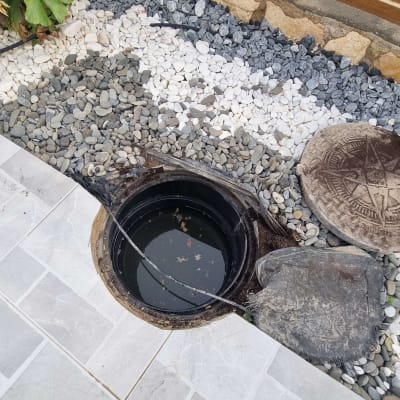 פתח ביוב פתוח עם מכסה בצד, מוקף באבנים קטנות