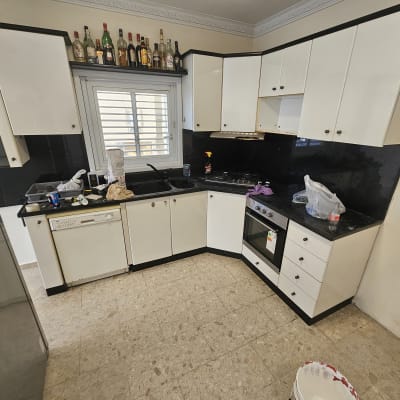 מטבח עם ארונות לבנים ומשטח עבודה שחור, בקבוקי שתייה על המדף