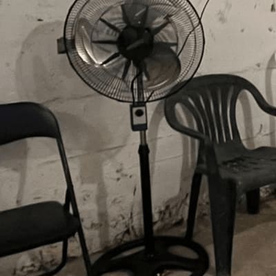 מאוורר עומד ליד כיסאות פלסטיק וקיר בטון