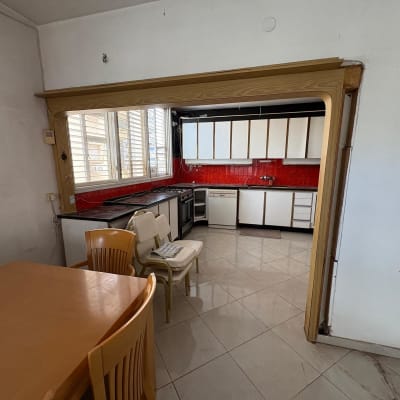 מטבח עם ארונות לבנים וקיר אדום, שולחן וכיסאות עץ.