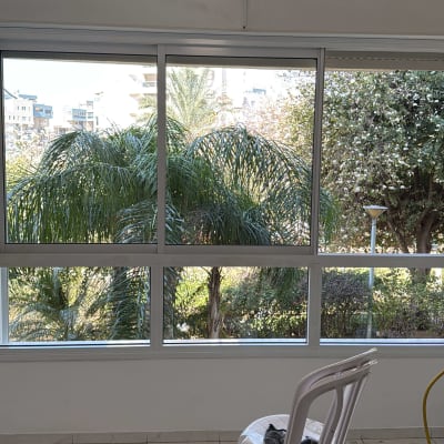 חלון גדול עם נוף לעצים, כיסא פלסטיק וצינור ניקוי על הרצפה.