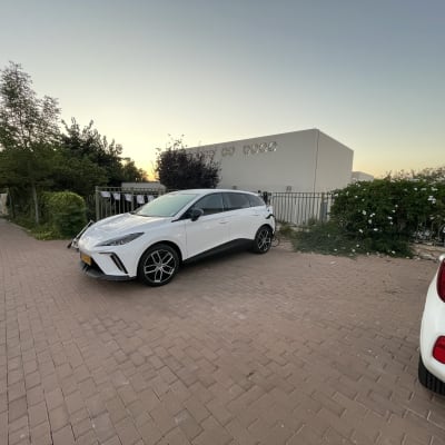 רכב לבן חונה ליד מבנה וגדר, עצים ברקע