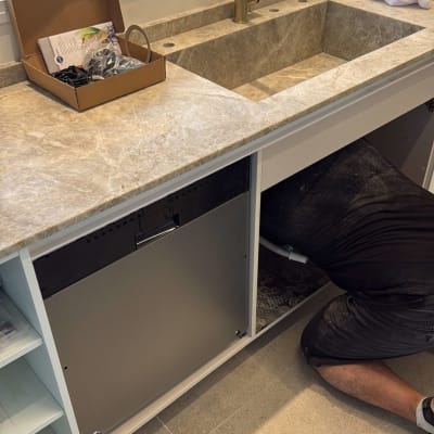 אדם עובד מתחת לכיור במטבח, כלים וציוד על השיש