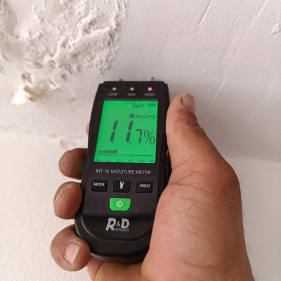 מד לחות מוחזק ביד ליד קיר עם רטיבות