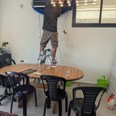 אדם מתקין מזגן על קיר בחדר עם שולחן וכיסאות.