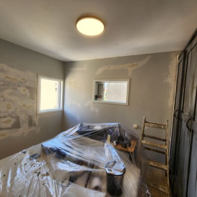 חדר עם קירות צבועים חלקית, סולם ויריעת פלסטיק על מיטה.