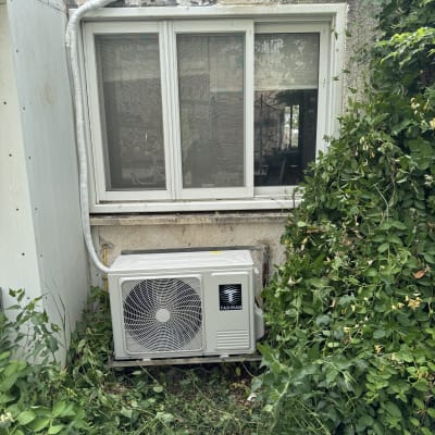 יחידת מזגן חיצונית מותקנת ליד חלון עם צמחייה מסביב