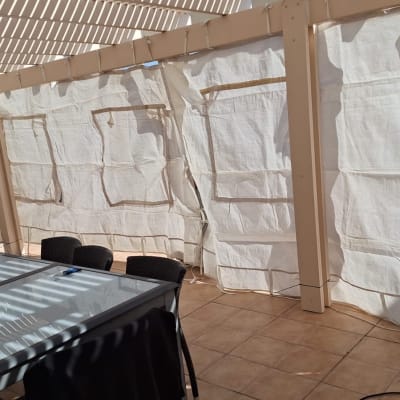 שולחן זכוכית וכיסאות תחת פרגולה עם וילונות לבנים