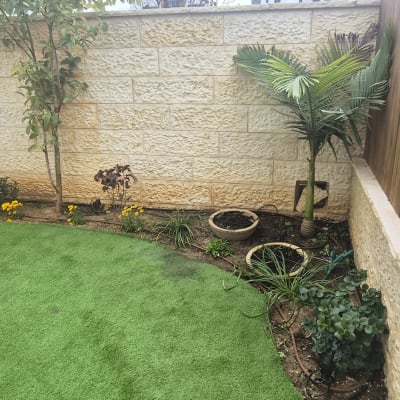 גינה עם דשא סינתטי, עצים ופרחים לאורך קיר אבן