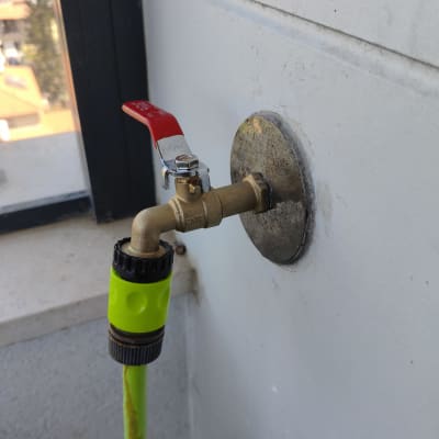 ברז מים חיצוני עם ידית אדומה וחיבור לצינור השקיה על קיר מרפסת