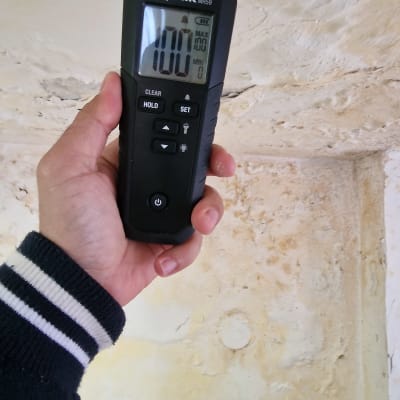 מד לחות מוחזק ביד מול קיר רטוב ומתקלף