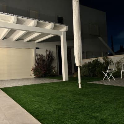 פרגולה לבנה בגינה עם דשא סינתטי וכיסאות פלסטיק לבנים