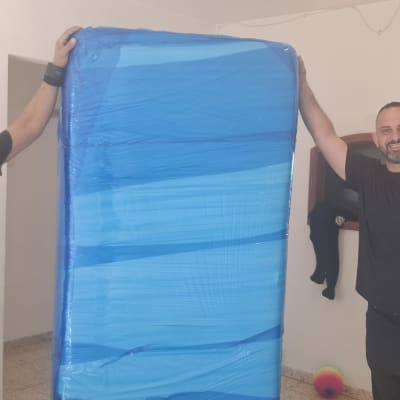 שני אנשים מחזיקים מזרן עטוף בניילון כחול בחדר ריק