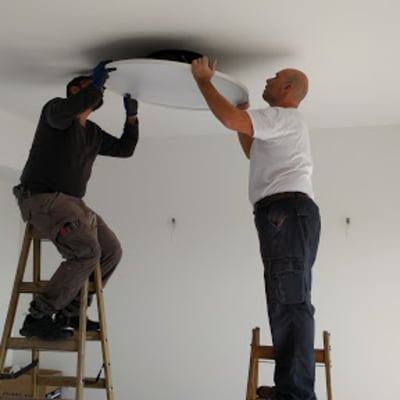 Two electricians installing a ceiling light fixture using ladders.