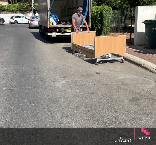 אדם מזיז מיטה על גלגלים ליד משאית פתוחה
