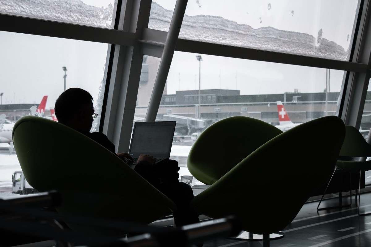 A traveller silhouetted against an airport window, working on a laptop.