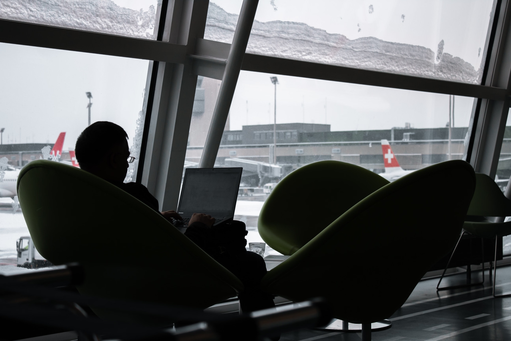 A traveller silhouetted against an airport window, working on a laptop.