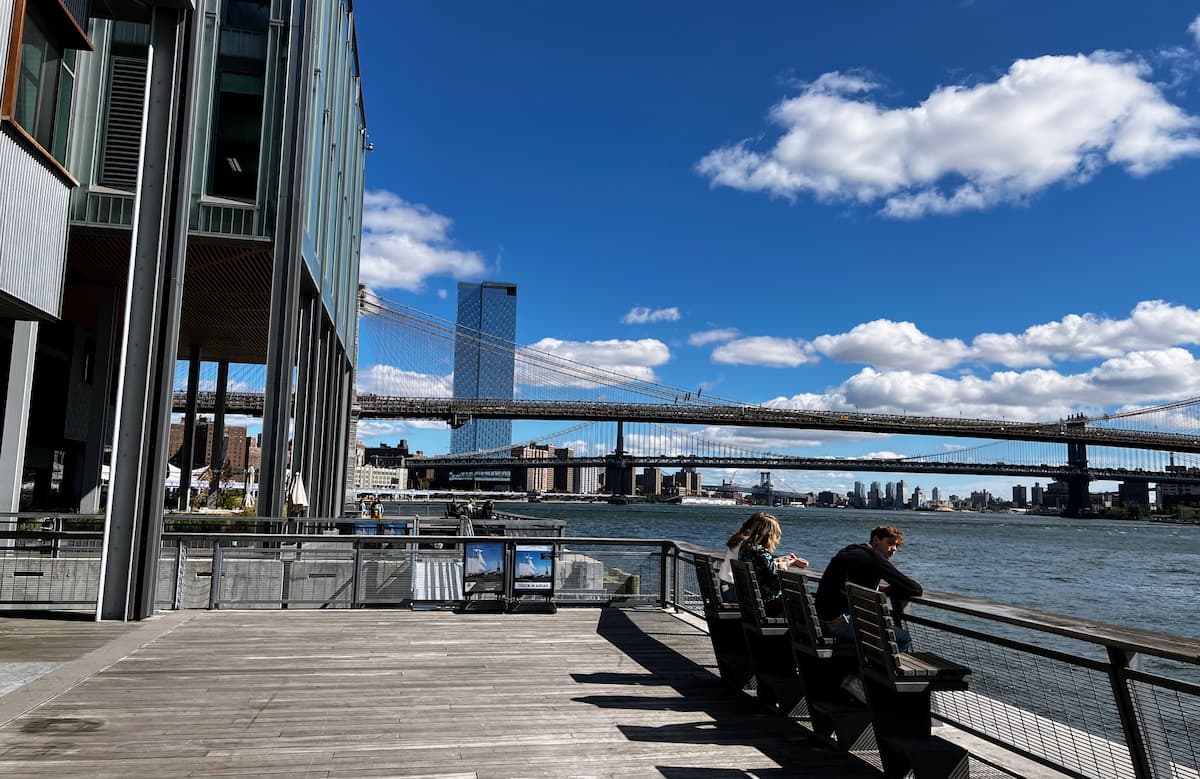 Photo of people sitting on benches overlooking a calm body of water, with two bridges and a tall building in the background