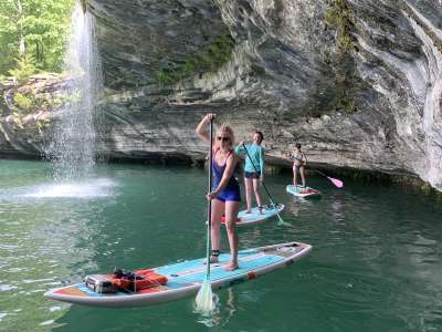 Three women standing up on light blue paddleboards. Water is below them. A limestone bluff on their left with a small waterfall in the background. Paddle Boarding to Waterfalls 