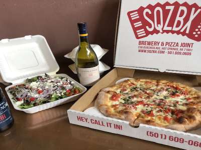 A to-go box of SQZBX pizza, salad and two bottles of wine sit on a table nicely arranged. 