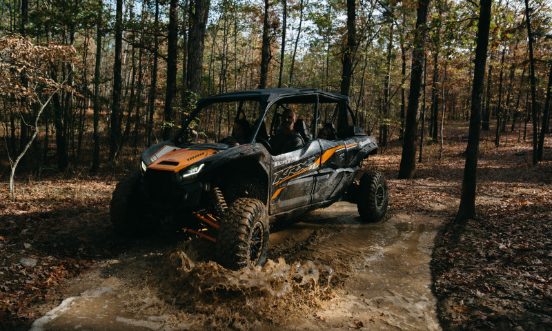 Orange and black side by side off road vehicle splashing through a mud puddle in a forest. 