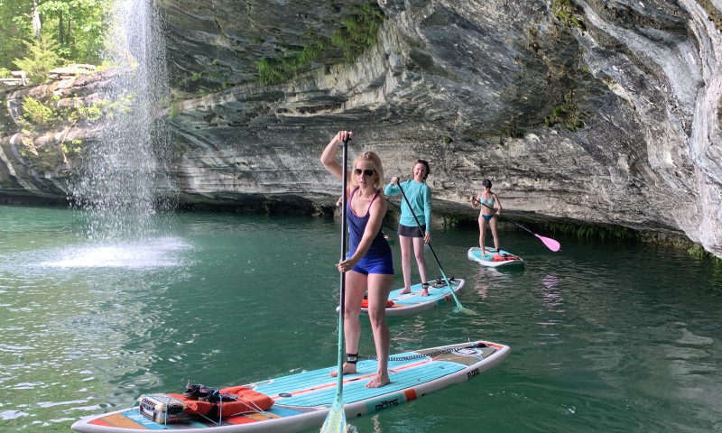 Three women standing up on light blue paddleboards. Water is below them. A limestone bluff on their left with a small waterfall in the background. Paddle Boarding to Waterfalls 