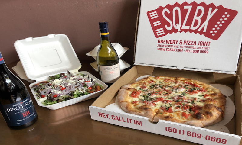 A to-go box of SQZBX pizza, salad and two bottles of wine sit on a table nicely arranged. 