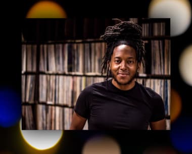 A person with dreadlocks smiles warmly, standing in front of a wall of vinyl records. The scene conveys a relaxed and inviting atmosphere.
