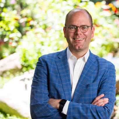 Isaac Thompson standing with arms folded, wearing blue suit and smiling in front of a white-green background.