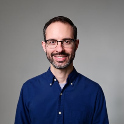 A headshot of David Stevens, wearing blue shirt, smiling and looking at camera in front of a grey background.
