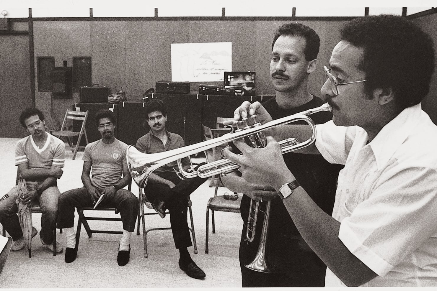 In a black and white photo, Manny Laureano plays the trumpet as a roomful of observers look on.