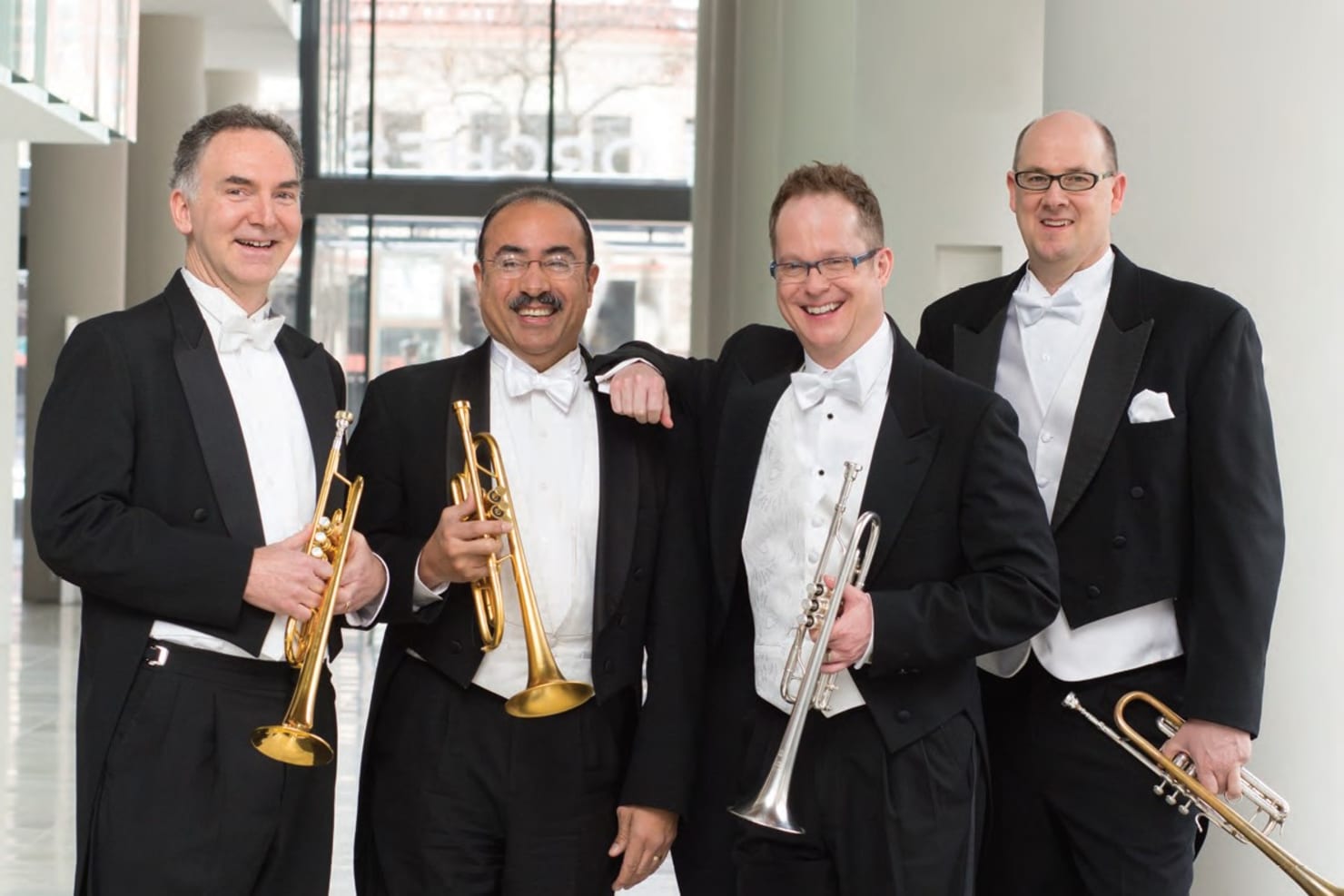 Four trumpet players standing and holding their trumpets, in formal concert dress in the Orchestra Hall lobby.