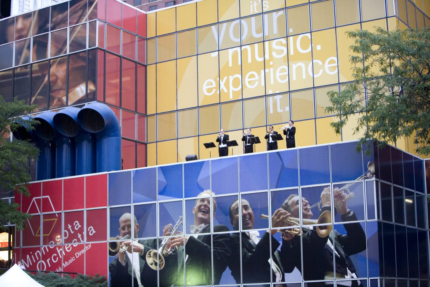 Four trumpet players stand on the rooftop of Orchestra Hall, as the Hall itself is decorated with a photo of the section.