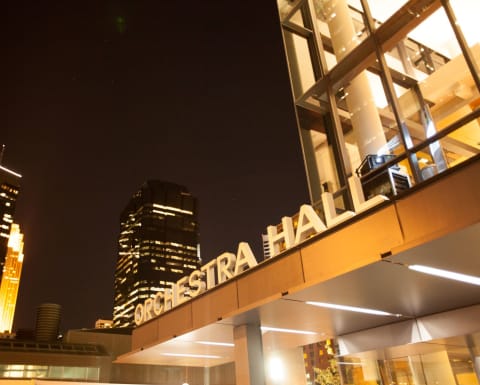 An exterior image of Orchestra Hall at night, with the downtown Minneapolis skyline in the background