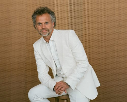 A portrait of Thomas Sondergard, posing in a white suit against a wood wall. He's leaning slightly forward while sitting on a stool.