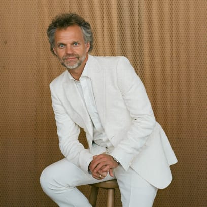 A portrait of Thomas Sondergard, posing in a white suit against a wood wall. He's leaning slightly forward while sitting on a stool.