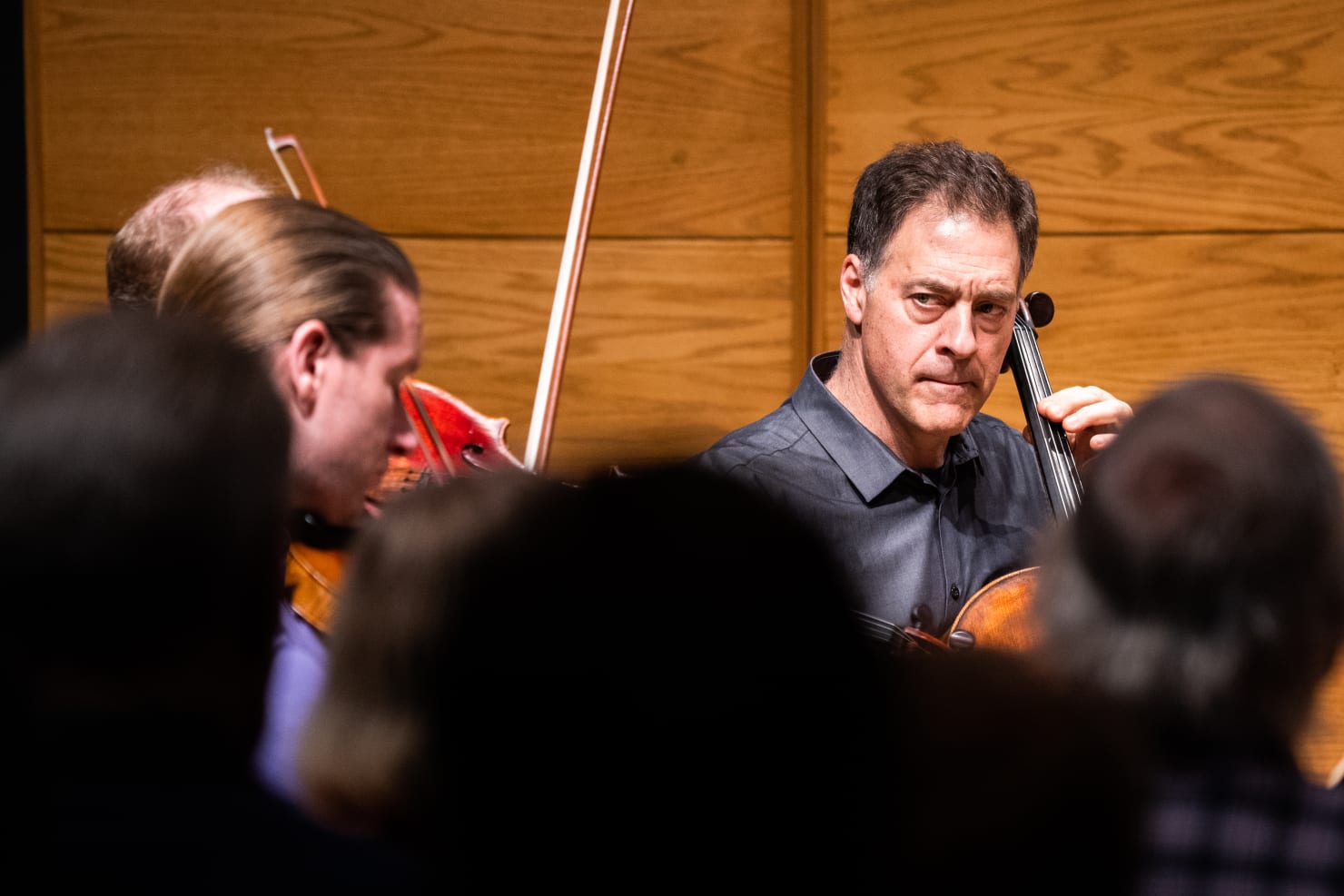 A close-up image of a violin player and cello player, the back of the heads of audience members visible and blurred in the image's foreground.