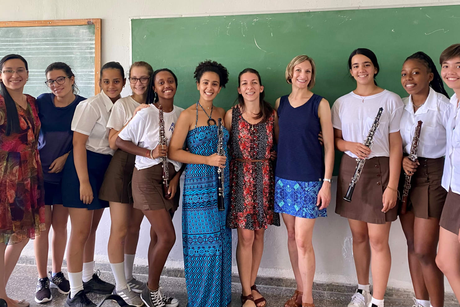 A group of students and two adult teachers standing together in a classroom in front of a chalkboard, posing with oboes and smiling at the camera.