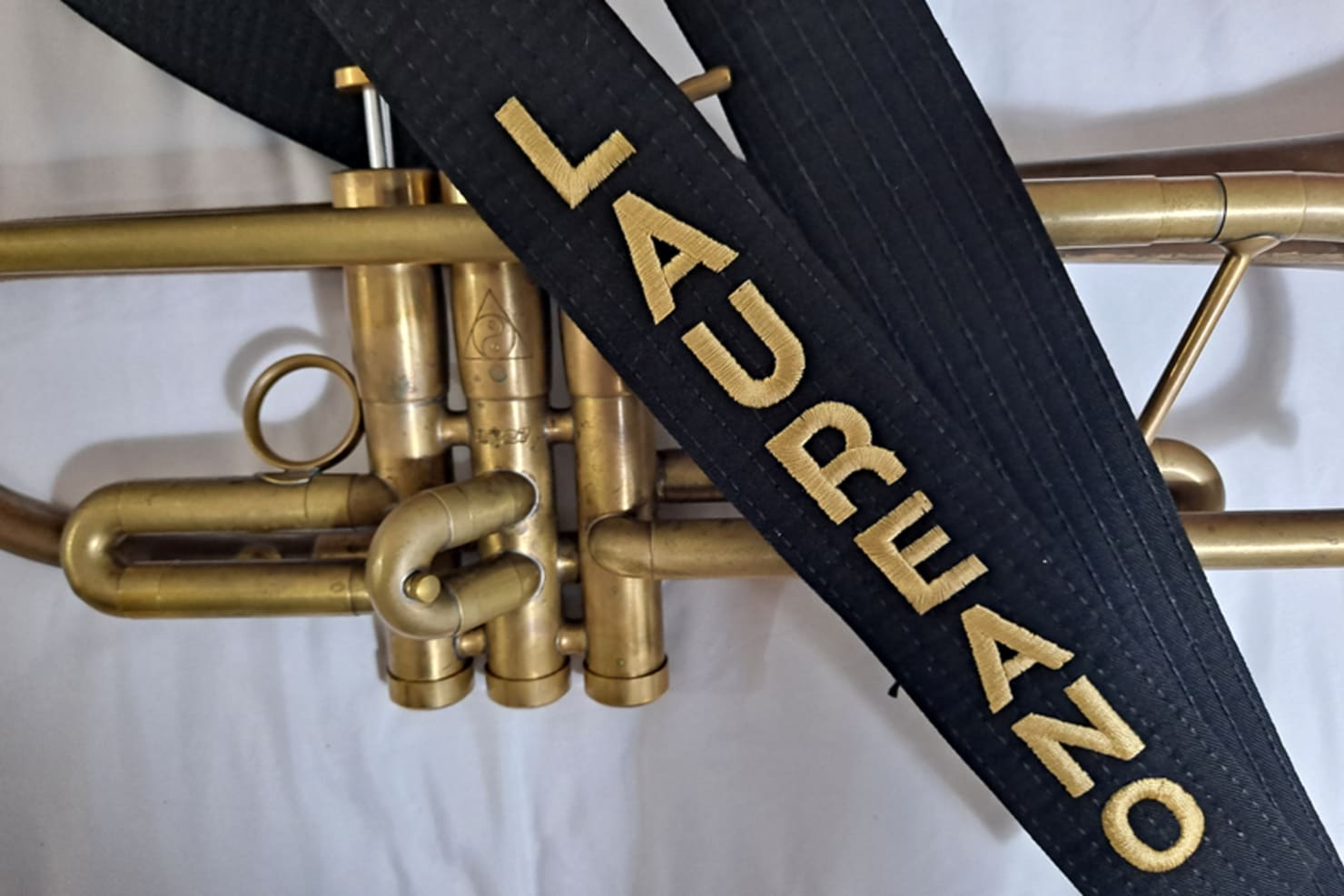 A close-up of a black belt with "LAUREANO" spelled out in gold letters; the belt is laid atop a trumpet.