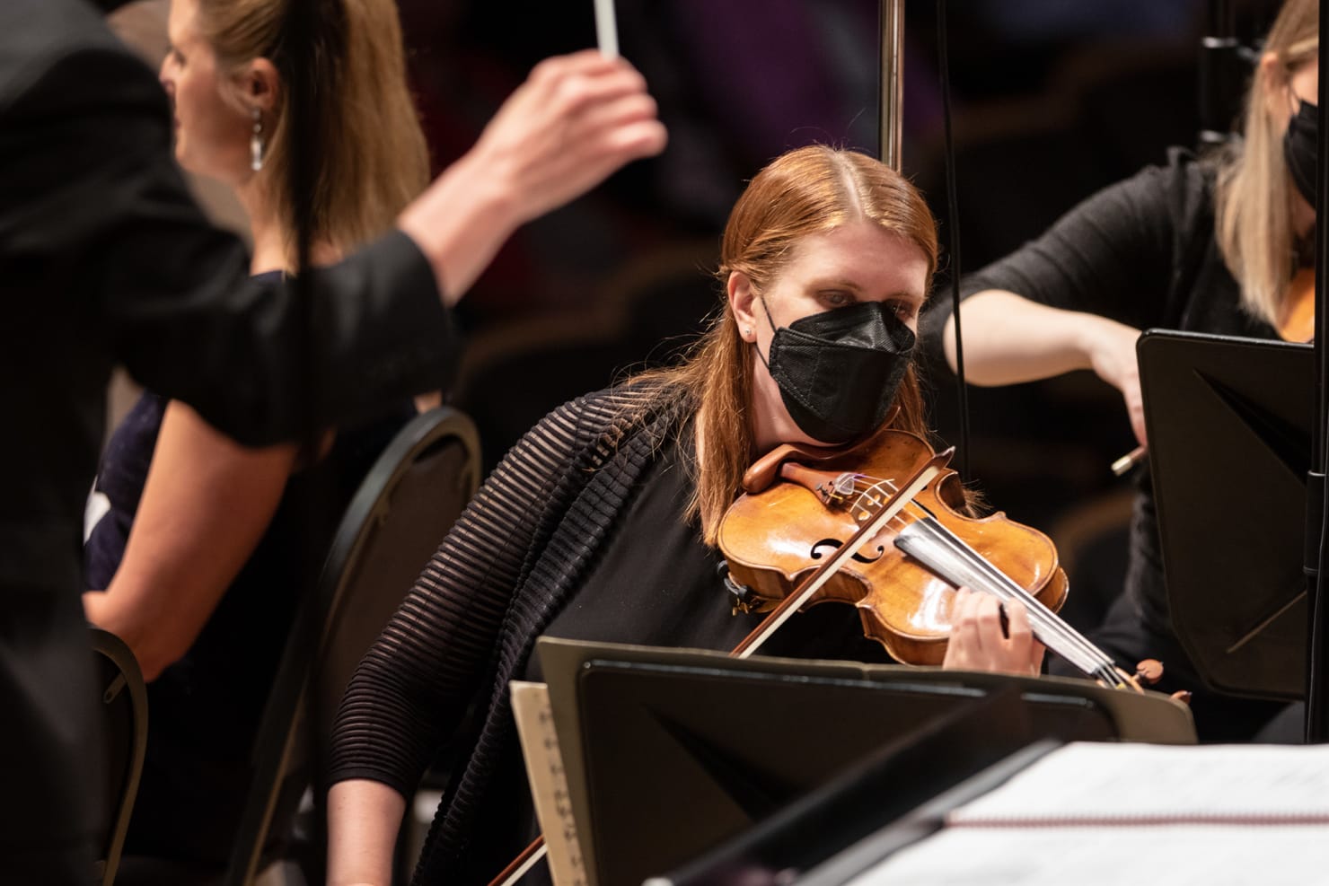 A close-up image of Erin Keefe playing the violin onstage during a concert, dressed in all-black with a black mask.