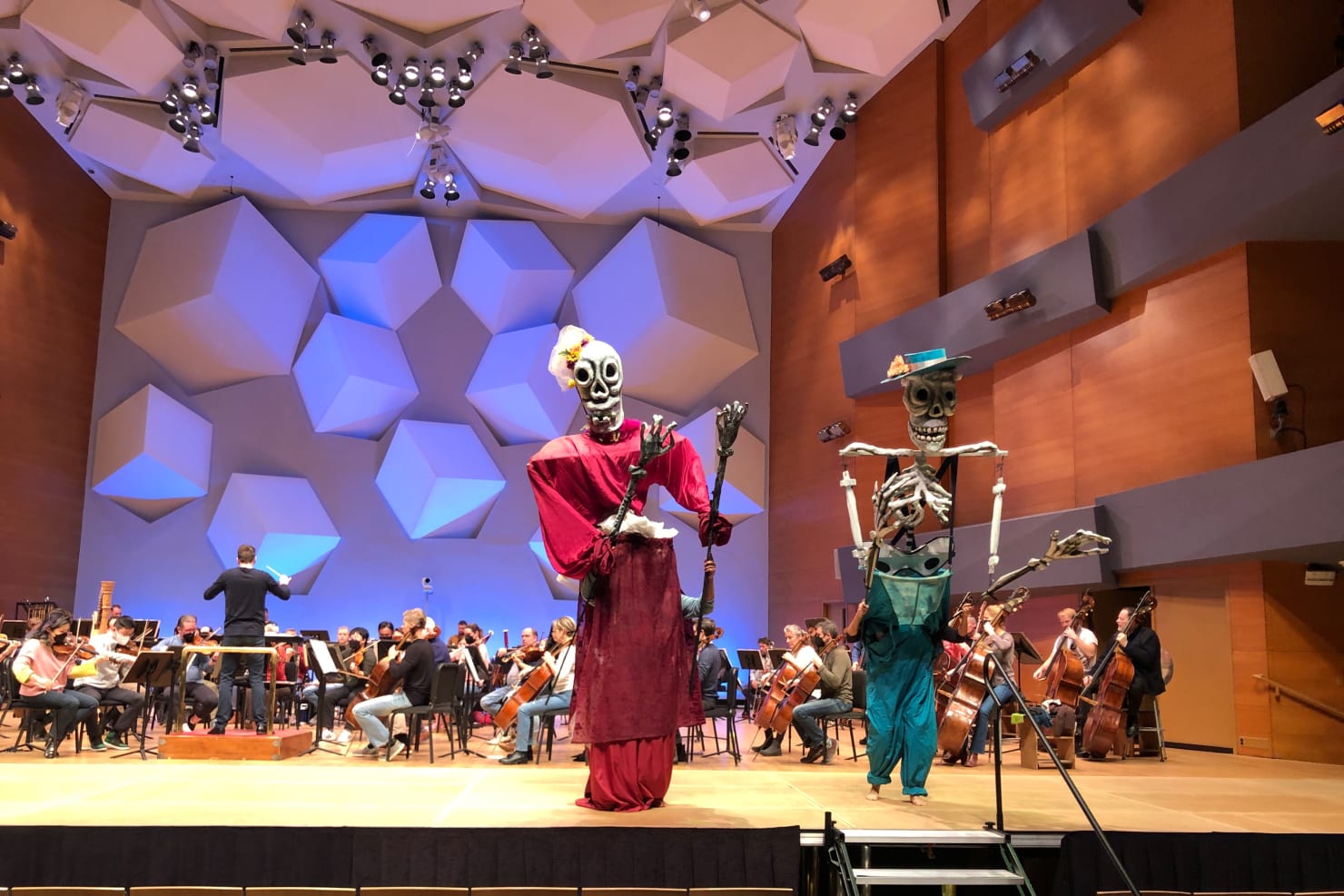 A scene from a rehearsal on the stage of Orchestra Hall; before the ensemble are two large skeleton puppets, one in red and the other in blue.